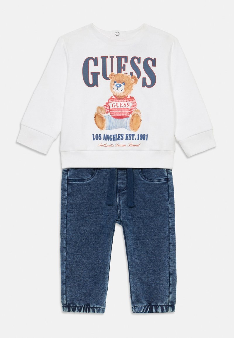 Weißer Pullover mit Bärenprint und "GUESS"-Logo, kombiniert mit dunkelblauen Jeans mit elastischem Bund und gekrempelten Säumen.