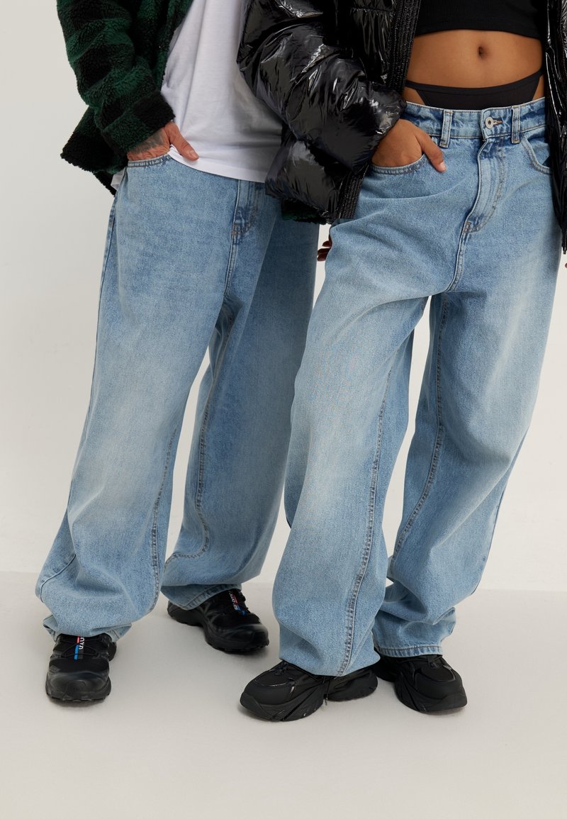 Ljusblå vida jeans i denim med en avslappnad passform och subtil blekning. Kombinerat med svarta sneakers, mot en neutral bakgrund.