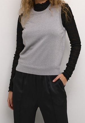 Femme portant un top argenté sans manches par-dessus un chemisier noir à manches longues texturé, avec un pantalon noir taille haute, une main dans la poche.