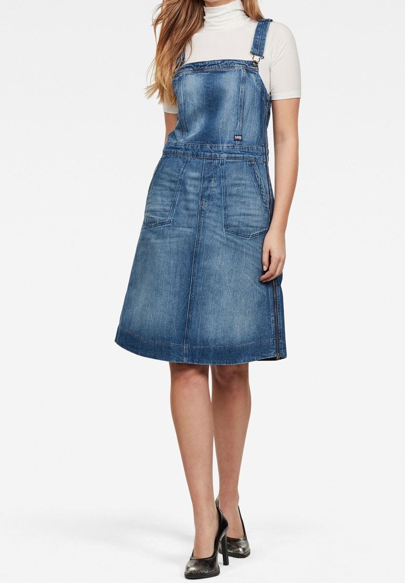 Femme portant une robe salopette en denim bleu avec poches avant et un haut à col roulé blanc à manches courtes, associée à des talons hauts noirs.