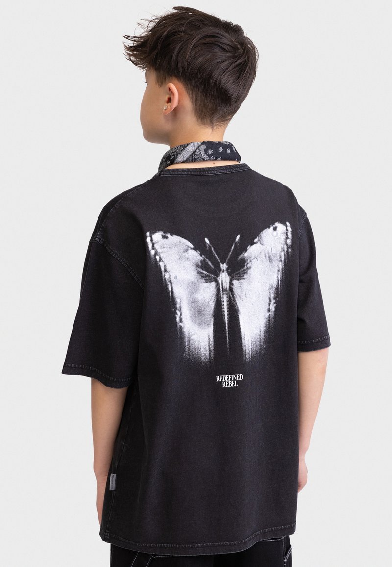 Schwarzes, übergroßes T-Shirt mit einem weißen Schmetterlingsgrafik auf dem Rücken und dem Text "REDEFINED REBEL" darunter. Weiche, denimartige Textur.