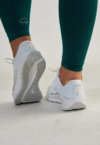 Białe buty sportowe z teksturowanym materiałem siatkowym, jasnoszare akcenty i wzorzysta podeszwa; noszone z zielonymi legginsami, które mają mały detal w kształcie serca.