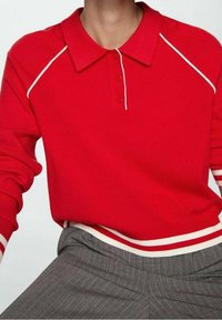 Person iført en rød langærmet polo-shirt med hvid kant og tre knapper, parret med grå stribede bukser.