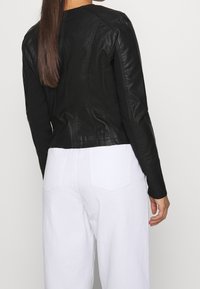 Giacca in pelle nera con design aderente, dettagli di cucitura visibili e maniche lunghe, abbinata a pantaloni bianchi a vita alta.