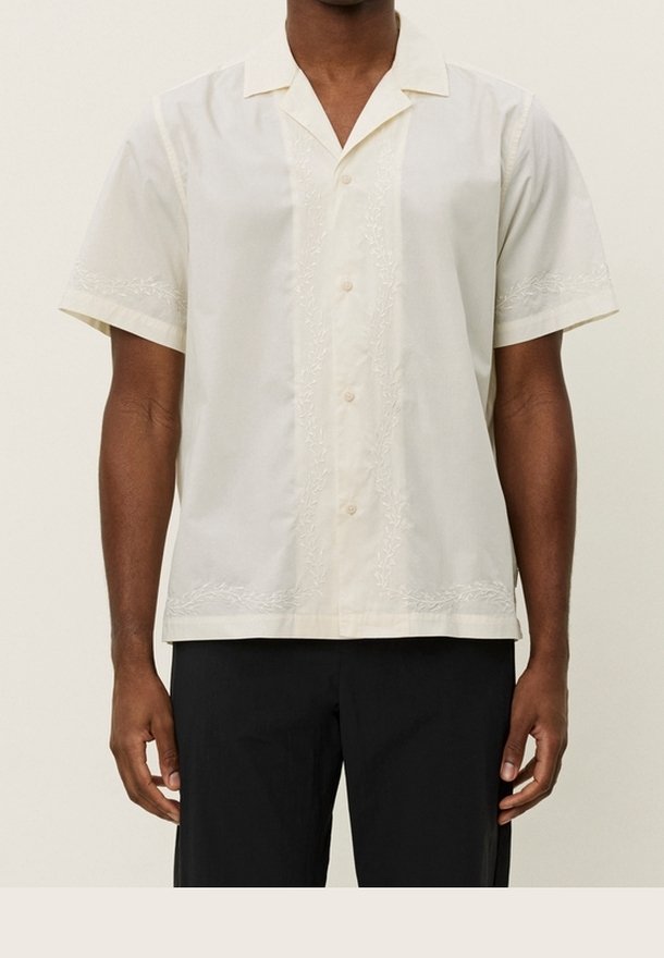 Homme portant une chemise crème à manches courtes avec des motifs brodés subtils et un pantalon noir, debout droit.