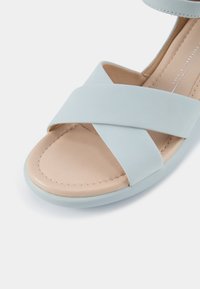 Vaaleansininen tasapohjainen sandaali, jossa on ristikkäiset hihnat, pehmeä nahkainen päällinen, vaaleanbeige pohja ja kontrastivärinen kumiulkopohja. Sileä pinta, jossa on näkyvät ompeleet.