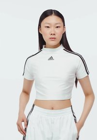 Biely skrátený športový dres s vysokým golierom a čiernymi tromi pruhmi. Na prednej strane sa nachádza logo Adidas a má priliehavý strih.