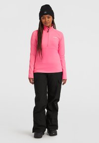 Top ajustado de color rosa, con manga larga y cremallera, y un alto collar, combinado con pantalones de esquí negros sueltos y botas de nieve negras. Tejido texturizado, diseño minimalista.