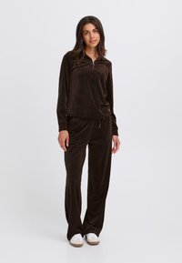 Bruin corduroy trainingspak met een half-rits top en broek met een trekkoord. Heeft brede benen en elastische boorden; gecombineerd met witte sneakers.