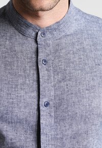 Camisa de lino azul de botonadura frontal con cuello mao, que cuenta con cuatro botones y una tela texturizada con un sutil patrón jaspeado.