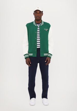 Groene en witte varsityjack met strepen, gedragen over een marinegestreept shirt, in combinatie met marineblauwe broek en witte sneakers. Witte pet op het hoofd.