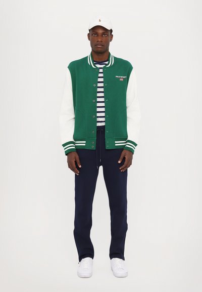 Giacca varsity verde e bianca con strisce, indossata su una camicia a righe blu, abbinata a pantaloni blu e sneakers bianche. Berretto bianco in testa.