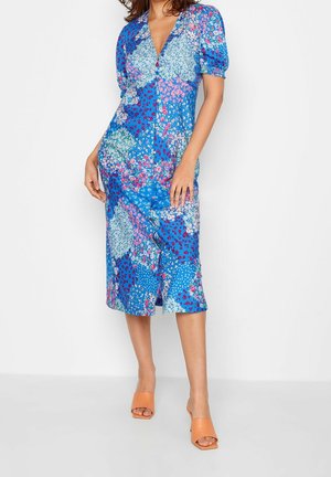 Vrouw die een blauwe midi-jurk met bloemenprint en pofmouwen draagt, met oranje open-toe pumps met hak, staand tegen een witte achtergrond.