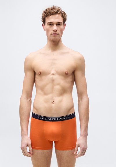 Νέος άνδρας με σγουρά μαλλιά, όρθιος, φορούντας πορτοκαλί boxer briefs Polo Ralph Lauren, σε απλό λευκό φόντο.