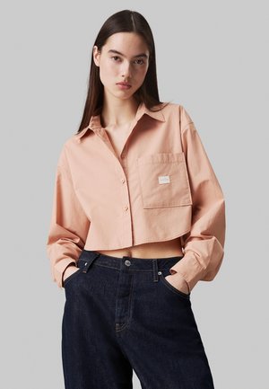 Cropped, light peach button-up shirt with a single breast pocket, paired with dark denim jeans. Cotton fabric with a smooth texture.