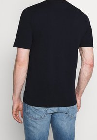 Muž v čistém černém tričku s krátkým rukávem a světle modrých džínách, stojící obrácený zády k jednobarevnému pozadí.