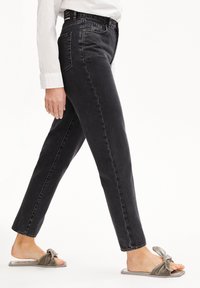 Svarta denimjeans med raka ben, med synlig sömnad och hög midja; kombineras med gråa slidessandaler med knutdetalj.