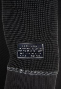 Nahaufnahme eines schwarzen, strukturierten Stoffärmel mit einem rechteckigen, marineblauen Etikett, das weißen gestickten Text zeigt und darunter eine detaillierte Naht hat.