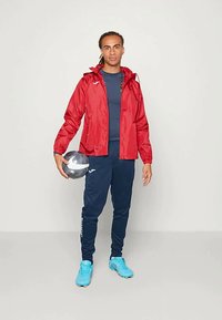 Junger Mann in roter Jacke, navyblauer Sportbekleidung und blauen Fußballschuhen, der einen schwarz-weißen Fußball hält und vor einem einfarbigen Hintergrund steht.