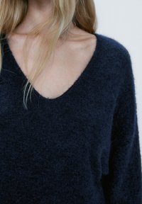 Lyshåret person iført en mørkeblå, fuzzy V-hals sweater, der viser nakke- og kraveområde mod en neutral baggrund.