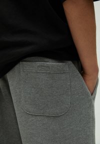 Pantalon de survêtement gris avec une texture douce, doté d'une poche arrière avec un détail de couture et d'une coupe décontractée. Porté avec un haut noir.