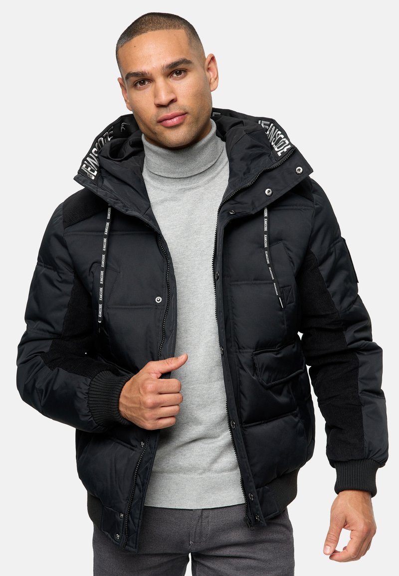 Chaqueta acolchada negra con capucha, que presenta mangas texturizadas en contraste, cordones ajustables, cierre frontal con botones a presión y múltiples bolsillos.