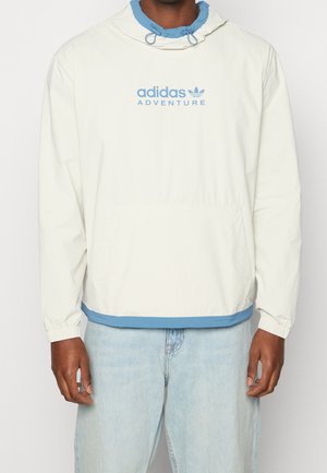Person trägt einen weißen Adidas Adventure-Pullover mit blauen Akzenten und hellblauen Jeans und steht mit entspannten Armen seitlich am Körper.