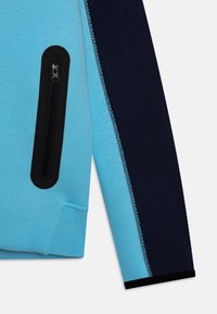 Vaaleansininen collegepaita tummansinisin yksityiskohdin, jonka tasku on vetoketjullinen ja kangas on teksturoitua. Suunnittelussa on kontrastiväriset hihat ja saumat.