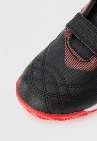 Scarpa sportiva nera con una parte superiore testurizzata, accenti rossi e una fascetta in Velcro. Presenta motivi cuciti e una suola in gomma.