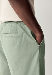 Shorts vert clair en tissu doux, avec une ceinture élastique, des poches latérales et une poche arrière avec un rabat rectangulaire.
