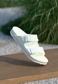 White sandal with two adjustable straps, made of smooth material, featuring a textured footbed and a lightly ridged sole.