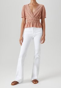 Pfirsichfarbige Bluse mit Puffärmeln, V-Ausschnitt und gerafftem Bund, kombiniert mit weißen Schlagjeans und braunen geflochtenen Sandalen.