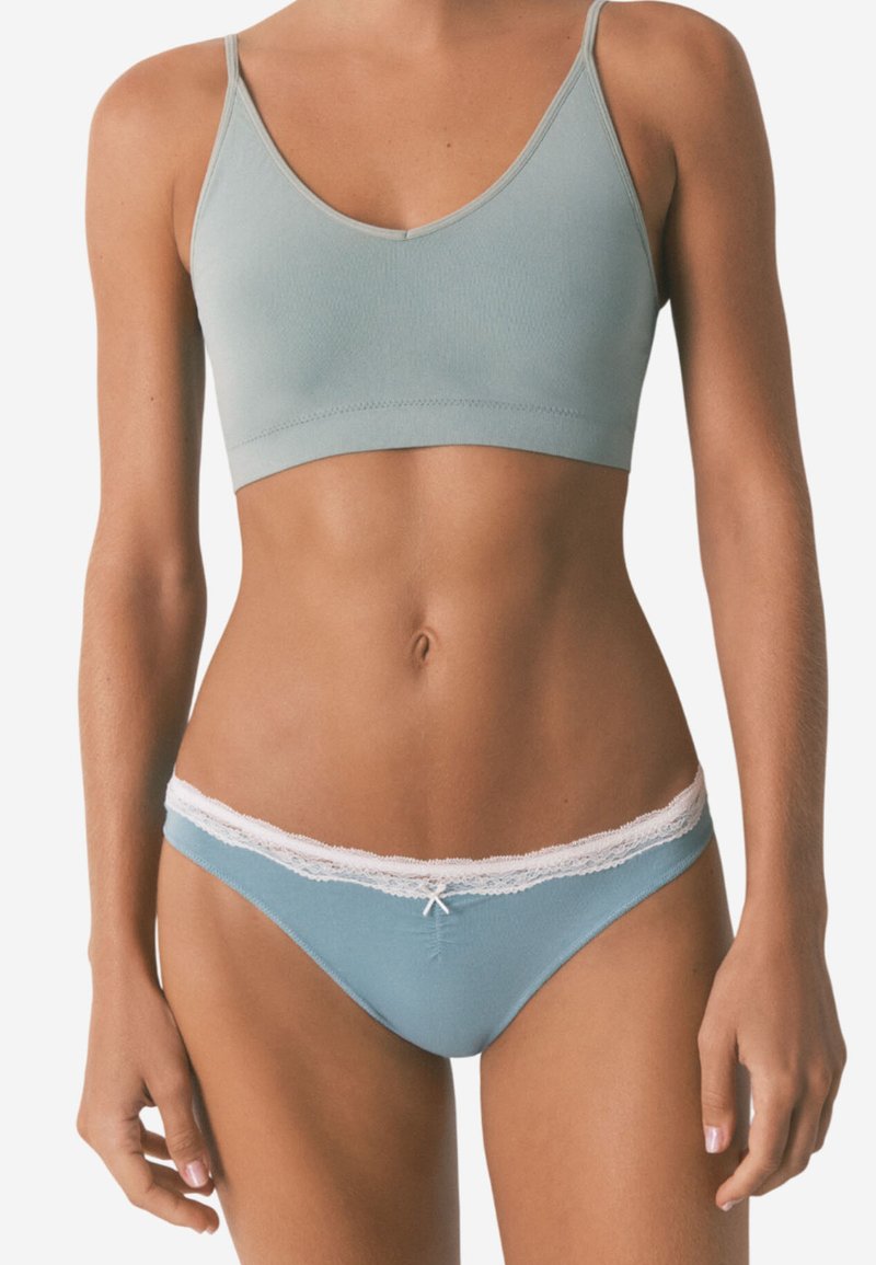 Donna che indossa un reggiseno senza cuciture di un azzurro chiaro e mutandine abbinate con pizzo, in piedi contro uno sfondo neutro.