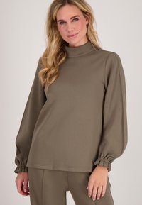 Vrouw in een olijfgroen langemouwen bovenstuk met gathered cuffs en bijpassende broek, staande tegen een effen lichte achtergrond.