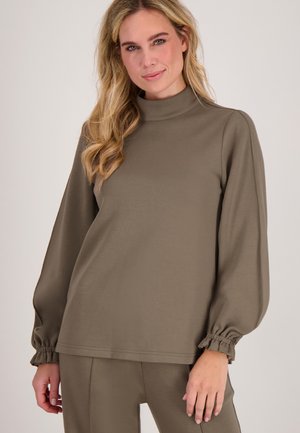 Vrouw in een olijfgroen langemouwen bovenstuk met gathered cuffs en bijpassende broek, staande tegen een effen lichte achtergrond.