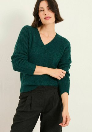 Türkiser Strickpullover mit V-Ausschnitt und geripptem Muster. Lange Ärmel; kombiniert mit dunkelkarierte Hosen, die ein lässiges Outfit zeigen.