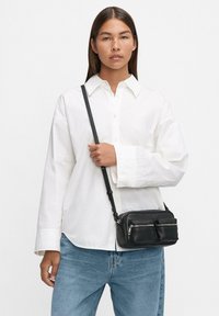Borsa a tracolla in pelle nera con due tasche con zip, abbinata a una camicia bianca con bottoni e jeans blu. Texture liscia, tracolla regolabile.