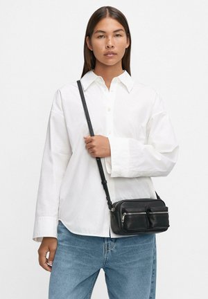 Borsa a tracolla in pelle nera con due tasche con zip, abbinata a una camicia bianca con bottoni e jeans blu. Texture liscia, tracolla regolabile.