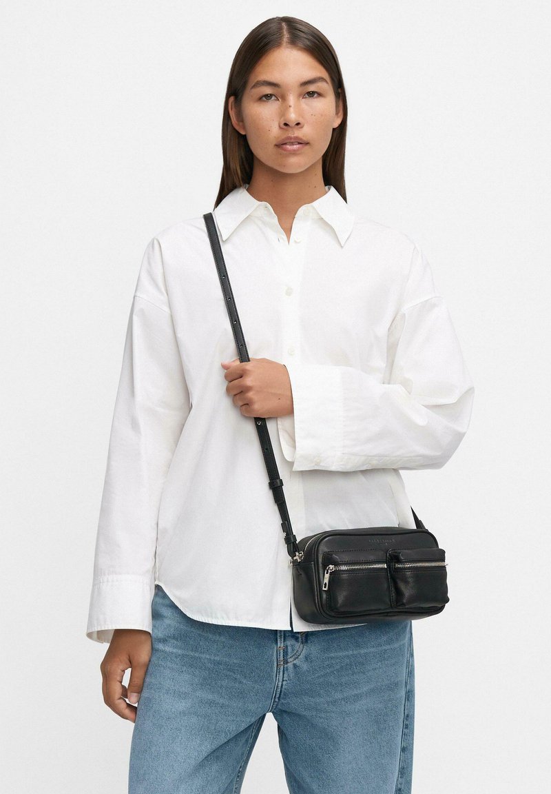 Borsa a tracolla in pelle nera con due tasche con zip, abbinata a una camicia bianca con bottoni e jeans blu. Texture liscia, tracolla regolabile.