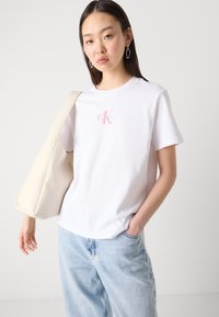Baltos medvilnės marškinėliai su trumpomis rankovėmis ir apvaliu iškirpimu, su šviesiai rožiniu ir mėlynu "Calvin Klein" logotipu krūtinėje.