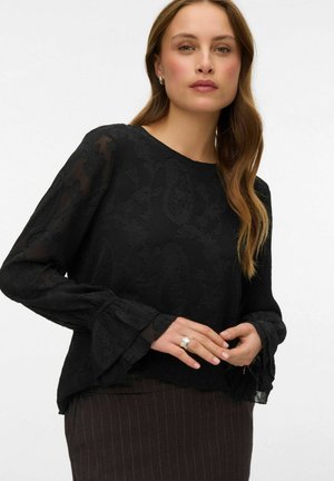 Sort bluse med blomsterprint, gennemsigtigt stof, rund halsudskæring og flagrende ærmer. Modellen har en ring på og en nederdel med nålestriber.