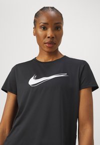 Must musta Nike särg lühikeste varrukatega, millel on rindade kohal silmapaistvalt valge swoosh logo, valmistatud sujuvast ja kerge kangast.