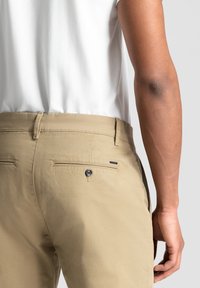 Pantaloni din bumbac bej cu un buzunar pe spate dotat cu un buton negru, cusături vizibile și un etichetă pe lateral. Modelul poartă o cămașă albă.