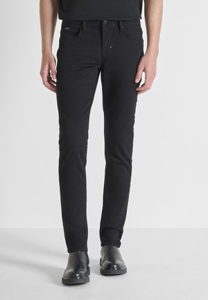Homme portant un jean noir slim avec des bottes en cuir noir, debout devant un fond gris clair uni.