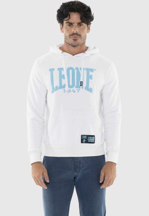 Uomo con capelli scuri che indossa una felpa bianca con cappuccio con la scritta "LEONE 1947" in blu e pantaloni blu scuro, in piedi contro uno sfondo semplice.