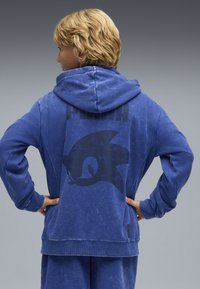 Felpa blu realizzata in morbido tessuto, con una grande grafica nera di un riccio e la parola "PUMA" sul retro. Inclusi maniche lunghe e cappuccio.