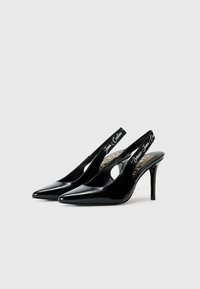 Black patent leather slingback heels feature a pointed toe and a cutout design, with a slim heel and contrasting white logo accents.