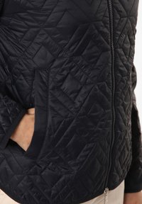 Steppjacke in Schwarz mit durchgehendem Reißverschluss, seitlichen Taschen und einem strukturierten Diamantmuster auf dem Stoff, mit glatter Oberfläche und körpernahem Schnitt.