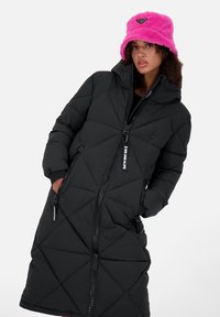Cappotto piumino trapuntato nero con cappuccio, dotato di zip frontale e tasche laterali. Indossato con un cappello a secchiello peloso di un brillante colore rosa.