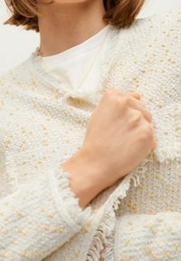 Frau, die einen strukturierten, cremefarbenen Pullover mit gelben Punkten und fransigen Kanten trägt, und den Stoff am Kragen über einem weißen Hemd hält.